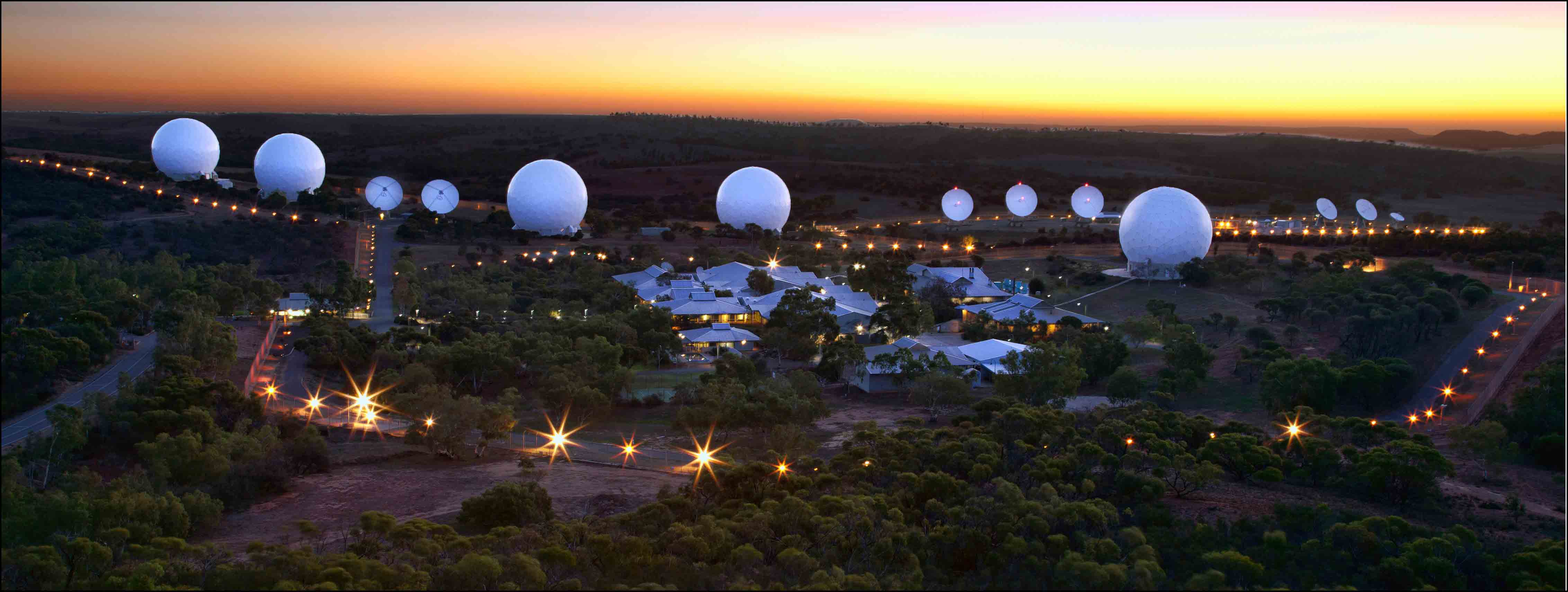 Lights, satellite dishes, buildings and large white spherical structures in a green landscape at dusk.