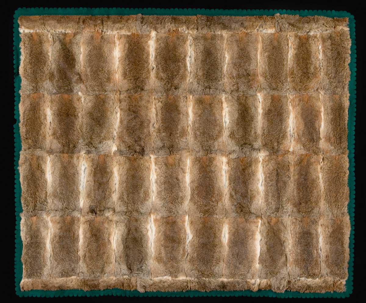 A large, brown coloured, rabbit skin rug that is lined and edged with emerald green felt. The felt edging is scalloped. There are 40 rectangular pelts used in the main section of the rug and several pelts cut into thinner strips that run along the two longer edges of the rug.