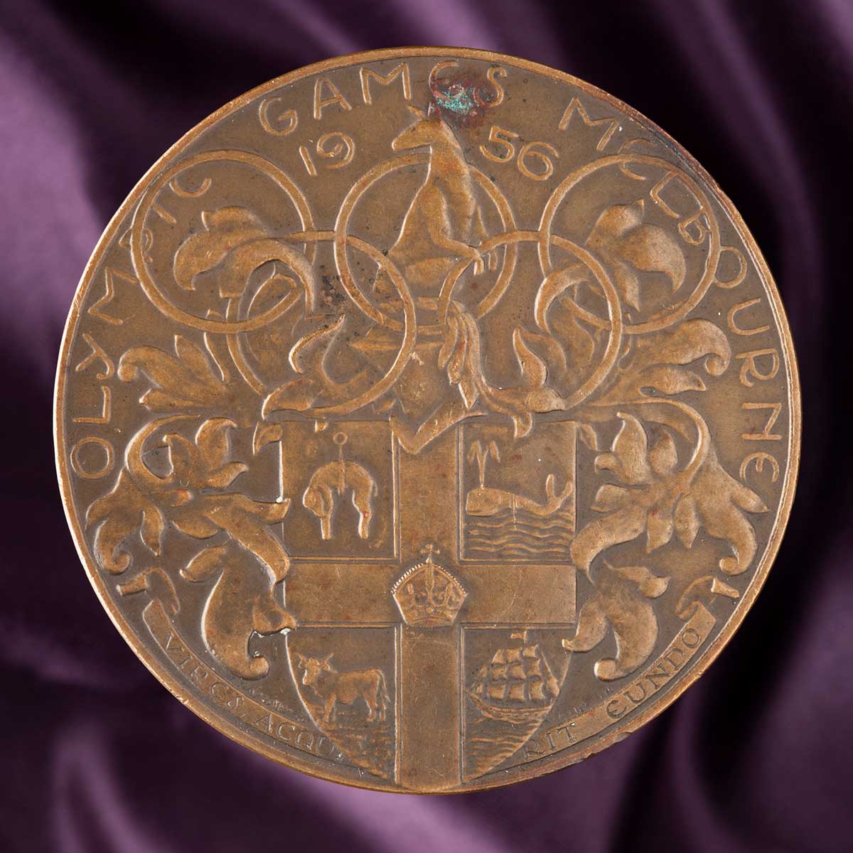 A circular bronze medal depicting a relief design of the Coat-of-Arms of the City of Melbourne surmounted by the five Olympic rings and bearing the inscription 'OLYMPIC GAMES MELBOURNE / 1956 / VIRES ACQU? [?] RIT EUNDO.' In addition, there is a floral design and a kangaroo positioned within the central ring.
