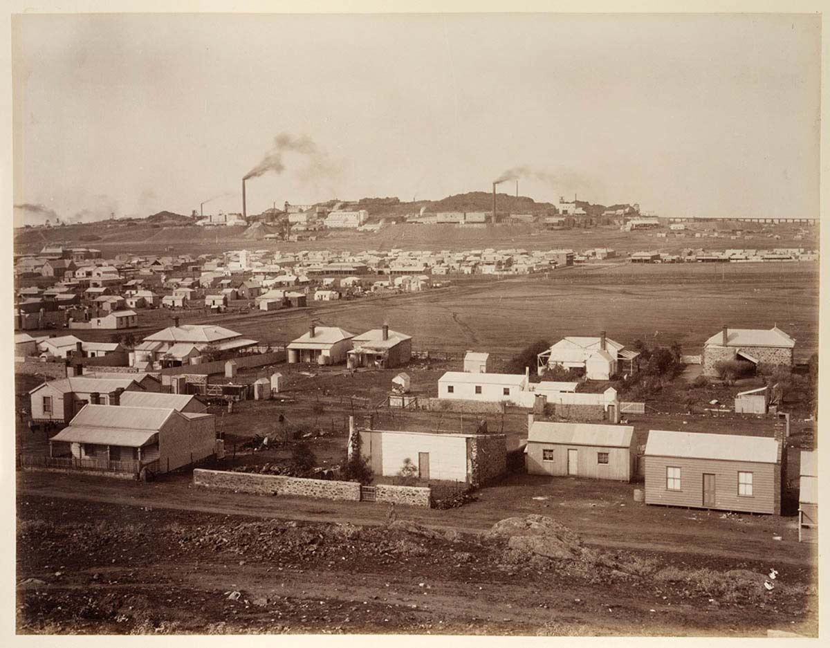 Black and white photograph of a township. In the distance a factory with smoke stacks can be seen.