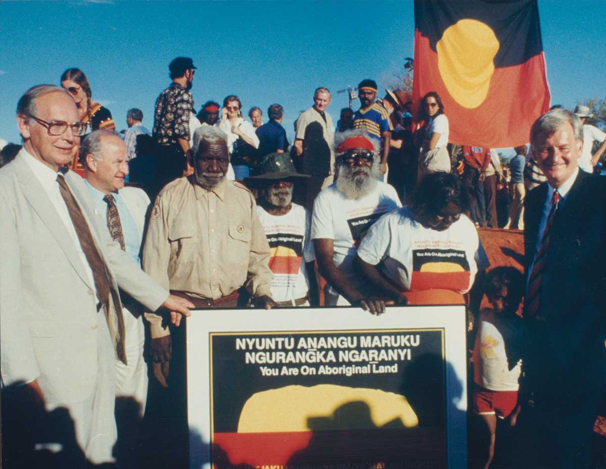 <p>Traditional owners Peter Bulla, Peter Kanari, Nipper Winmarti and his wife, Barbara Tjirkadu, with Sir Ninian Stephen and Clyde Holding (left) and Barry Cohen (right) and the special poster marking the handback of title to Ulu<u>r</u>u-Kata Tju<u>t</u>a National Park, 26 October 1985</p>