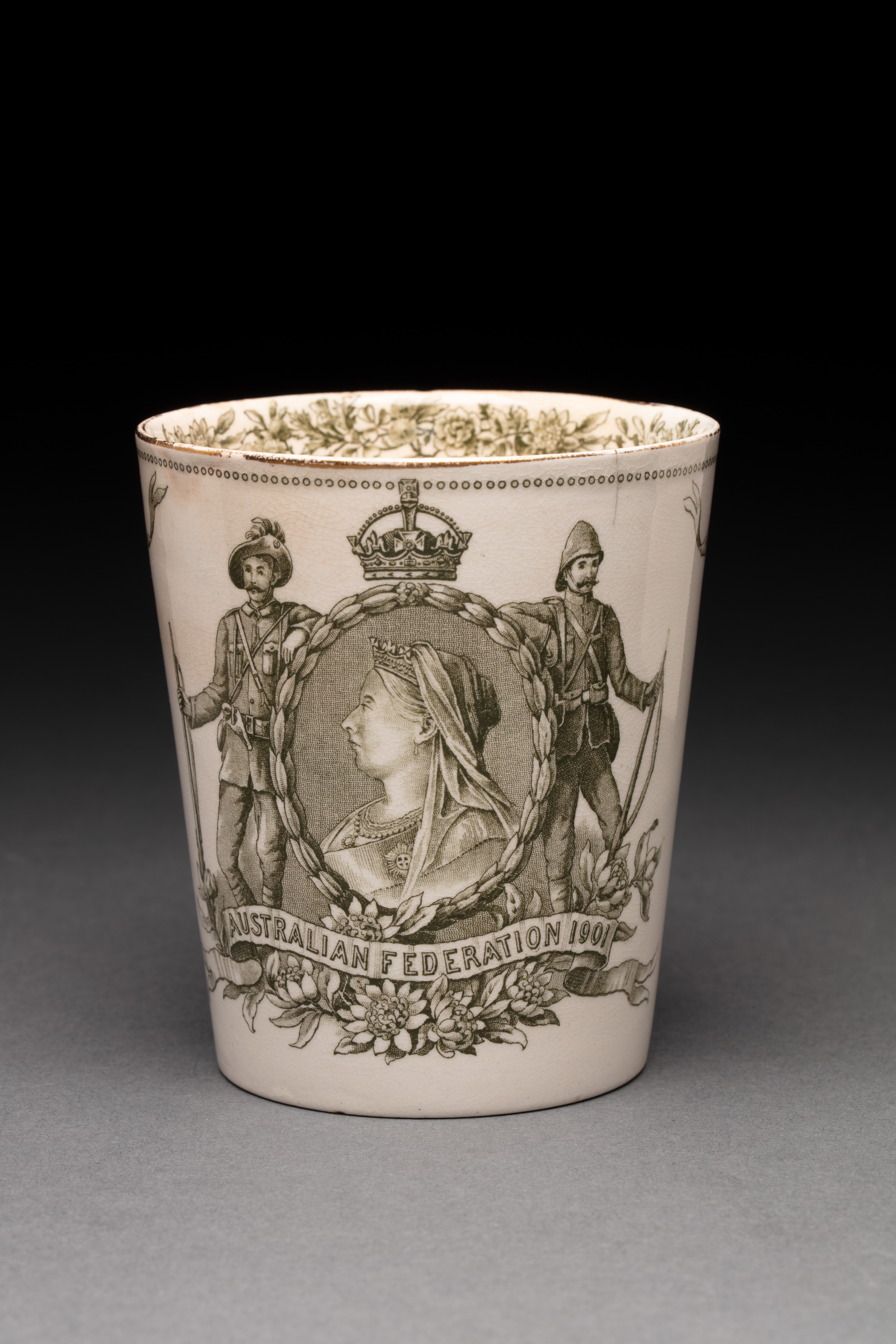  An earthenware beaker with a gilt rim. Decorated with a garland of flowers inside the rim, and on the front with a green transfer print of Queen Victoria centred inside a wreath border flanked by a British and an Australian soldier, and 'AUSTRALIAN FEDERATION 1901' inscribed within a ribbon amongst waratahs at the bottom. Printed portraits of Sir Henry Parkes and Lord Hopetoun are featured on the other side of the beaker between smaller decorative portraits of the Duke and Duchess of Cornwall and York.