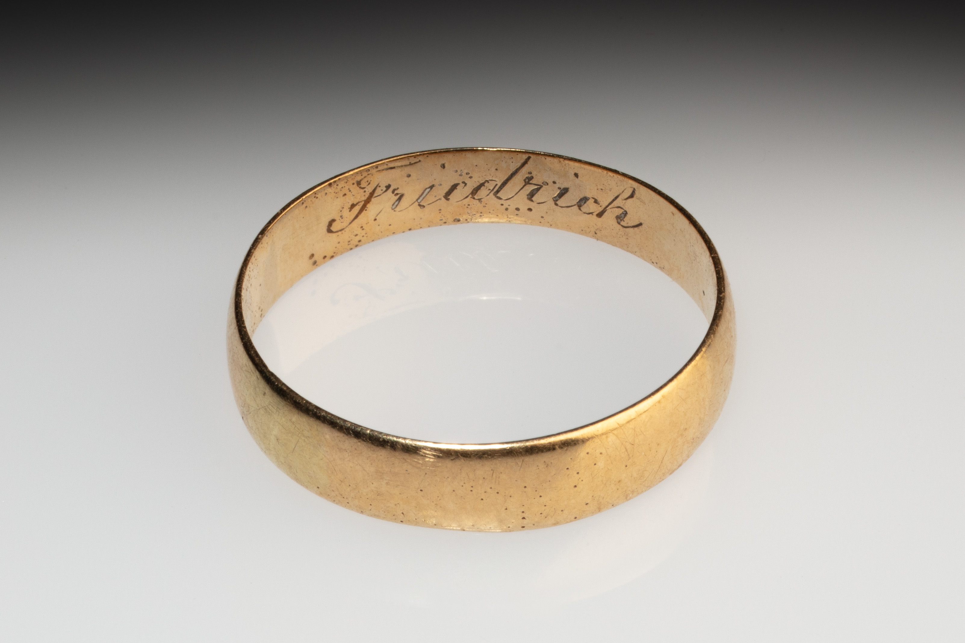 A gold wedding ring, engraved on the internal surface with the name 'Friedrich'. The ring is a plain gold band, with a slightly curved outer surface, and fine edges.