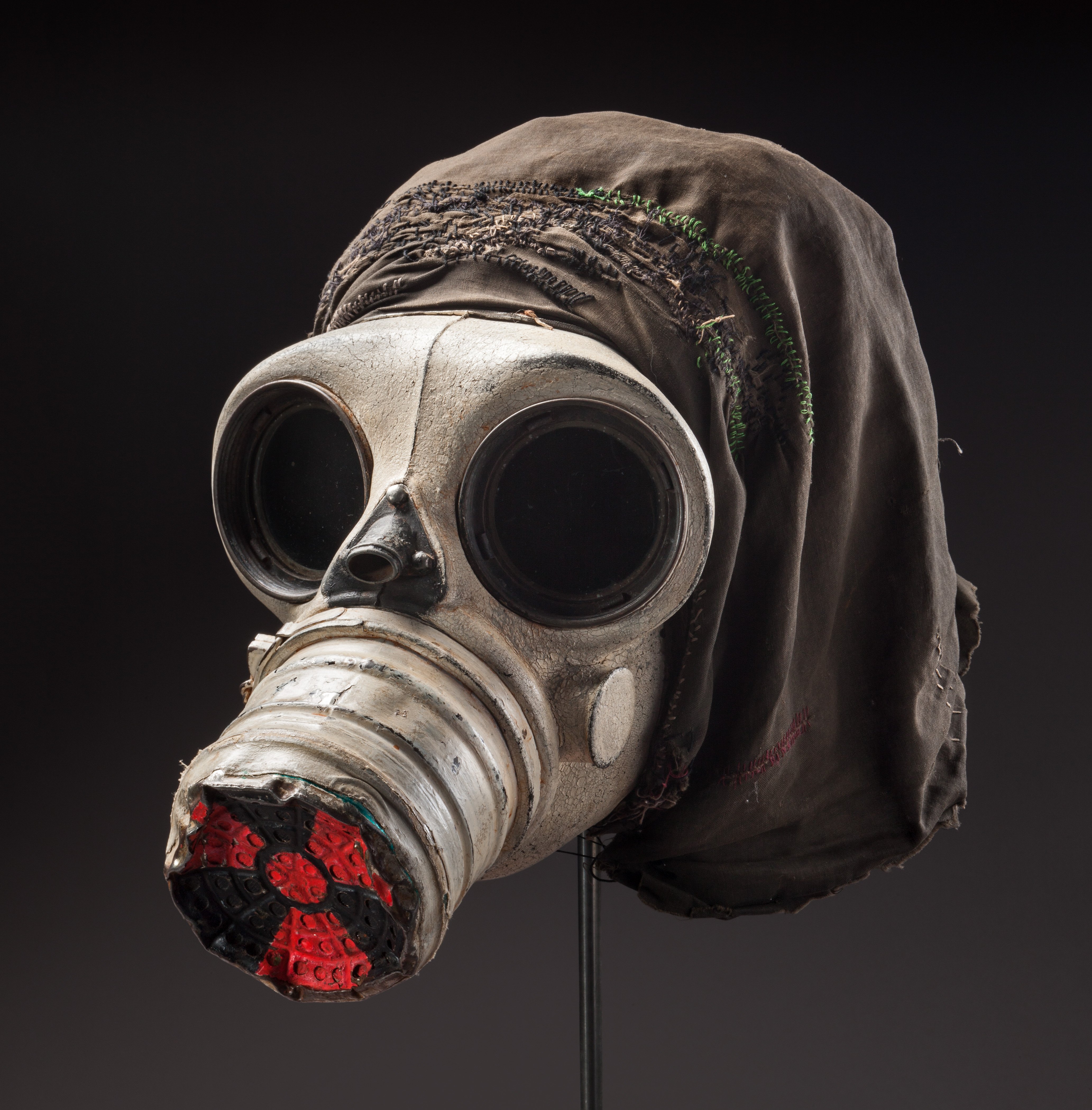  Gas mask painted red, white and black with black cloth hood attached. The front section has been painted white to give the appearance of a skull. The end of the nose cone has been painted red and black in the form of a radiation symbol. A black cloth hood has been sewn onto the back of the mask in order to fully cover the head when worn.