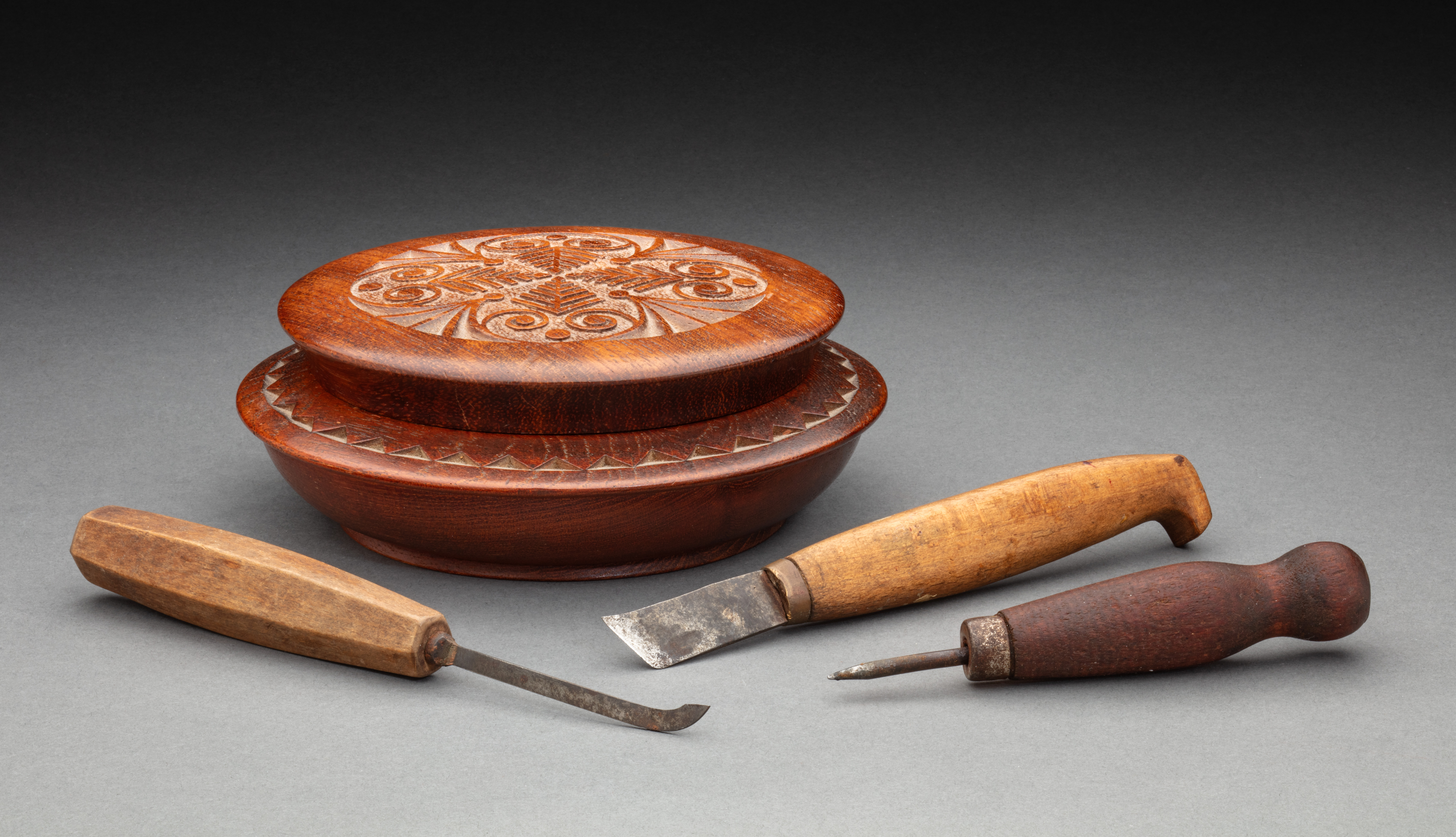  A shallow round carved wooden bowl with a removable lid. The bowl has a wide rim with a carved zig-zag pattern running around it. The lid sits above the bowl and has a short flange. On the top are carved decorations featuring curved and straight lines. The base is marked 'LATVIAN / D.P. WORK'.