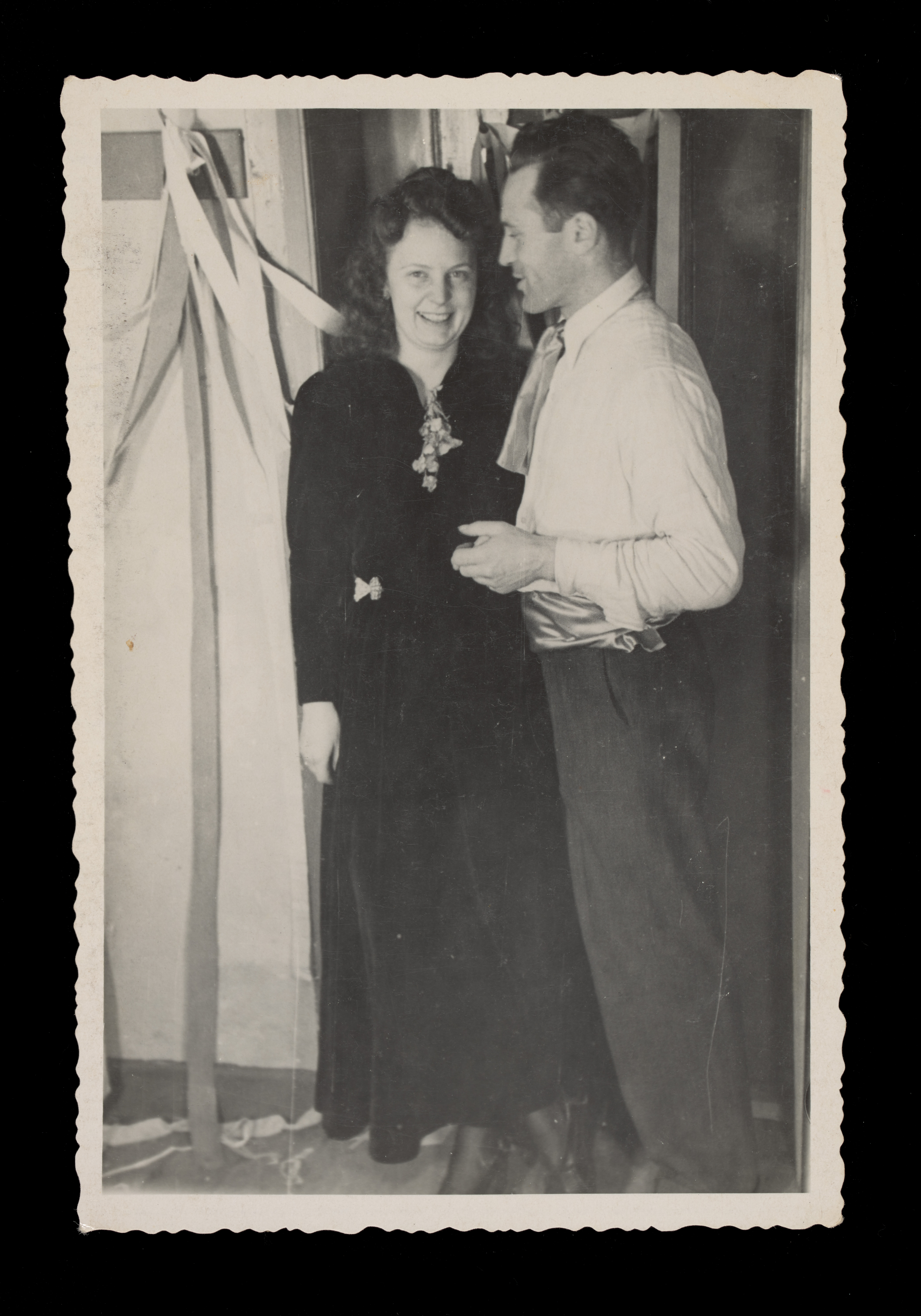  A black and white photograph of a man and woman standing near a wall. The man is standing sideways facing the woman and smiling. The woman is looking at the camera smiling. They appear to be indoors with ribbon like decorations behind them. The image has a plain border with a wavy edge. On the back in graphite pencil is the text 'Kinne' and '20'.