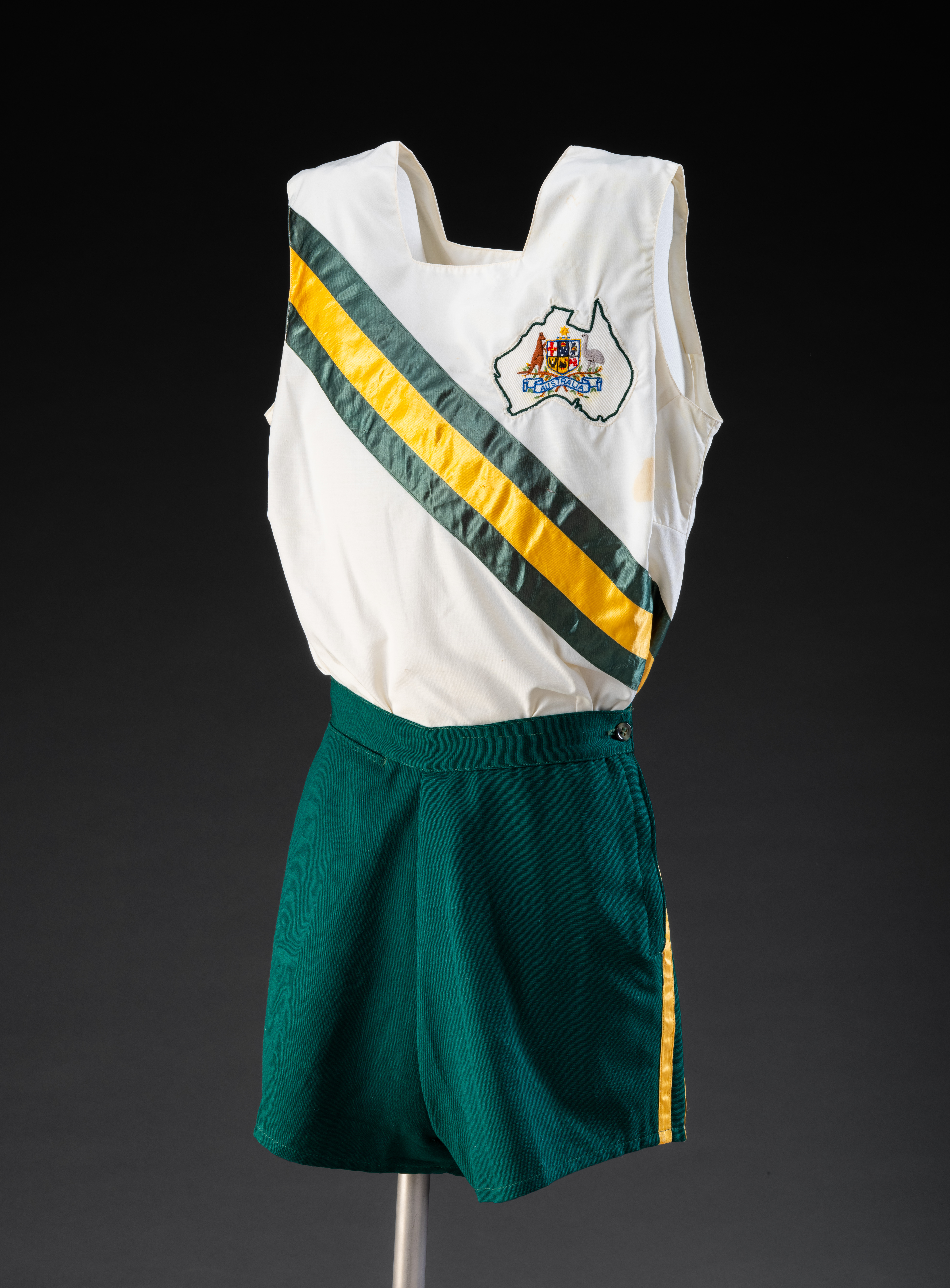  Australian Olympic Team athlete's white running singlet. It has diagonal green-gold-green stripes running from the right shoulder to the left hip, and an embroidered white badge on the left breast that depicts the Australian Coat-of-Arms inside a black outline of Australia. '1956 AUSTRALIAN / OLYMPIC TEAM / Official Garment / MADE BY / MELBOURNE SPORTS DEPOT' is on the label inside. It has a small stain on the front between the badge and the stripes. This singlet belonged to Shirley Strickland.