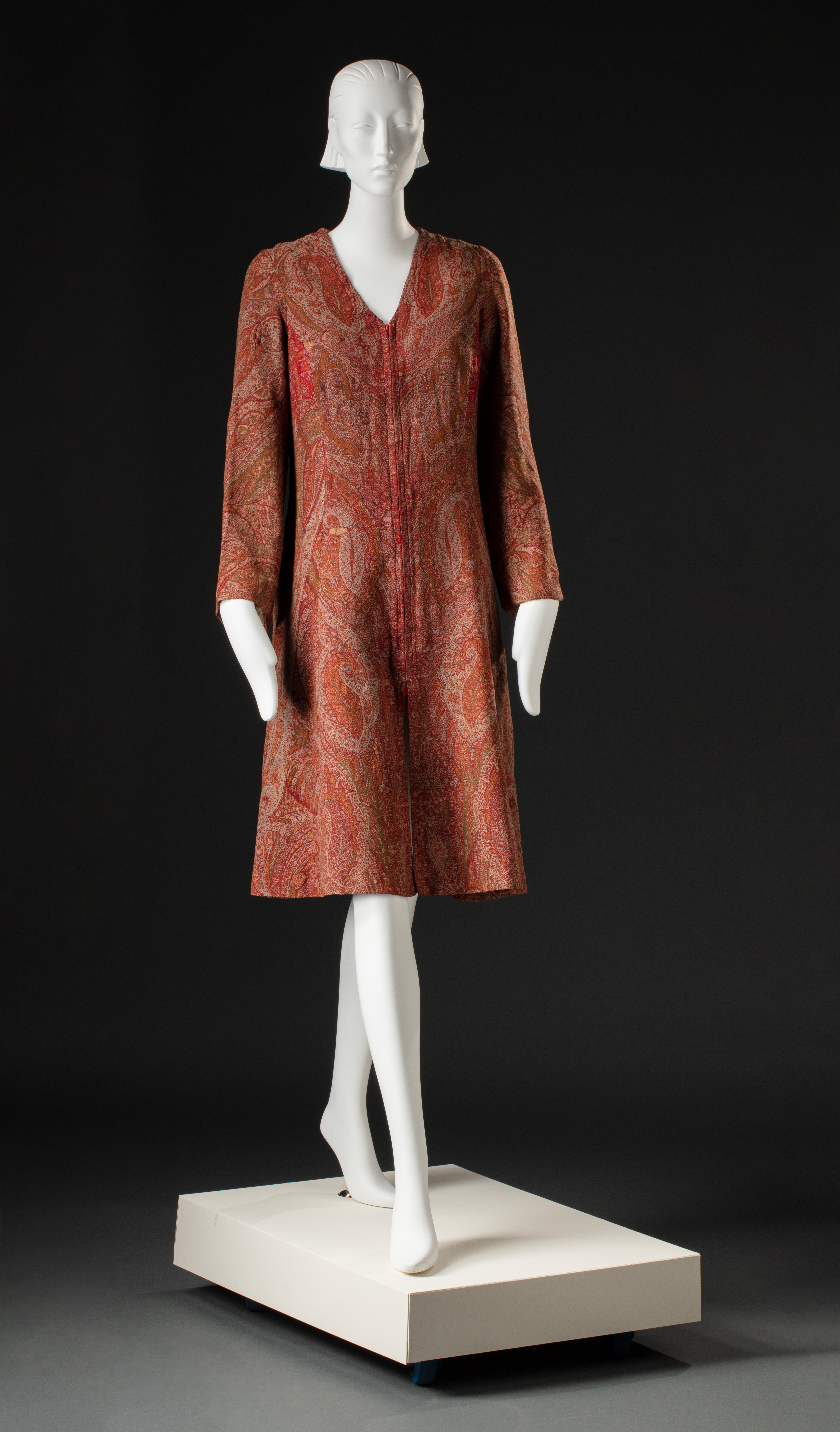 A handmade coat created from the fabric of a Paisley shawl. The A-line woollen coat has long sleeves and a V neck with a red zip sewn into the centre front. It is lined with a light brown viyella (wool and cotton blend fabric). The woven Paisley fabric is highly patterned and coloured light green, mid green, cream, orange, red and brown, and features a repeated teardrop motif. There are a number of small holes and tears in the fabric.