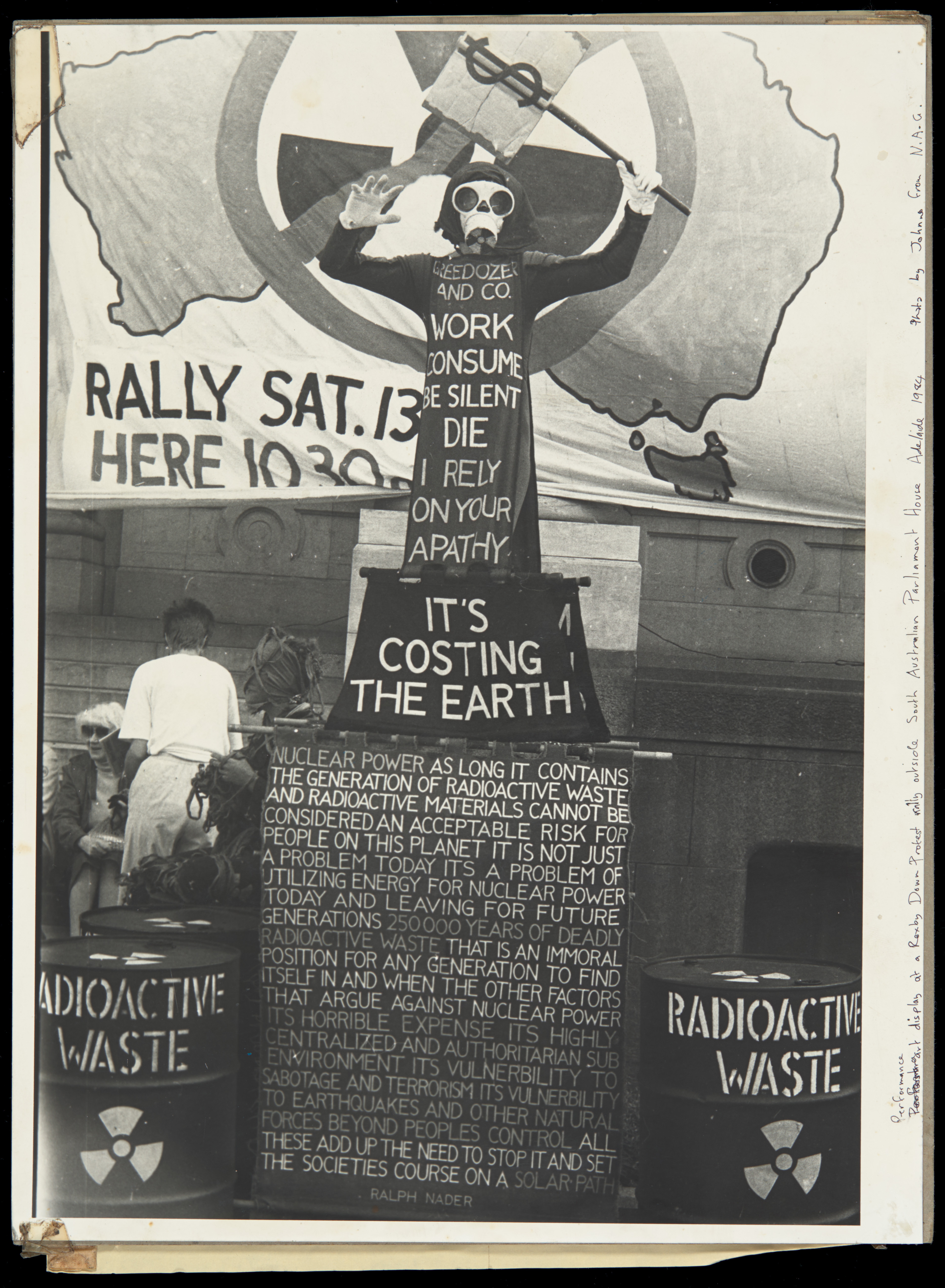  A photograph of Benny Zable in costume outside of the South Australian Parliament House in 1987. Zable is standing in front of a banner of Australia with a no nuclear sign. Handwriting is apparent on the side of the photo stating: "Performance and Display at a Roxby Downs Protest Rally outside South Australian Parliament House Adelaide 1984". The photograph is taped to a brown card with a "Peace Arts Victoria 86" pamphlet taped to the back.