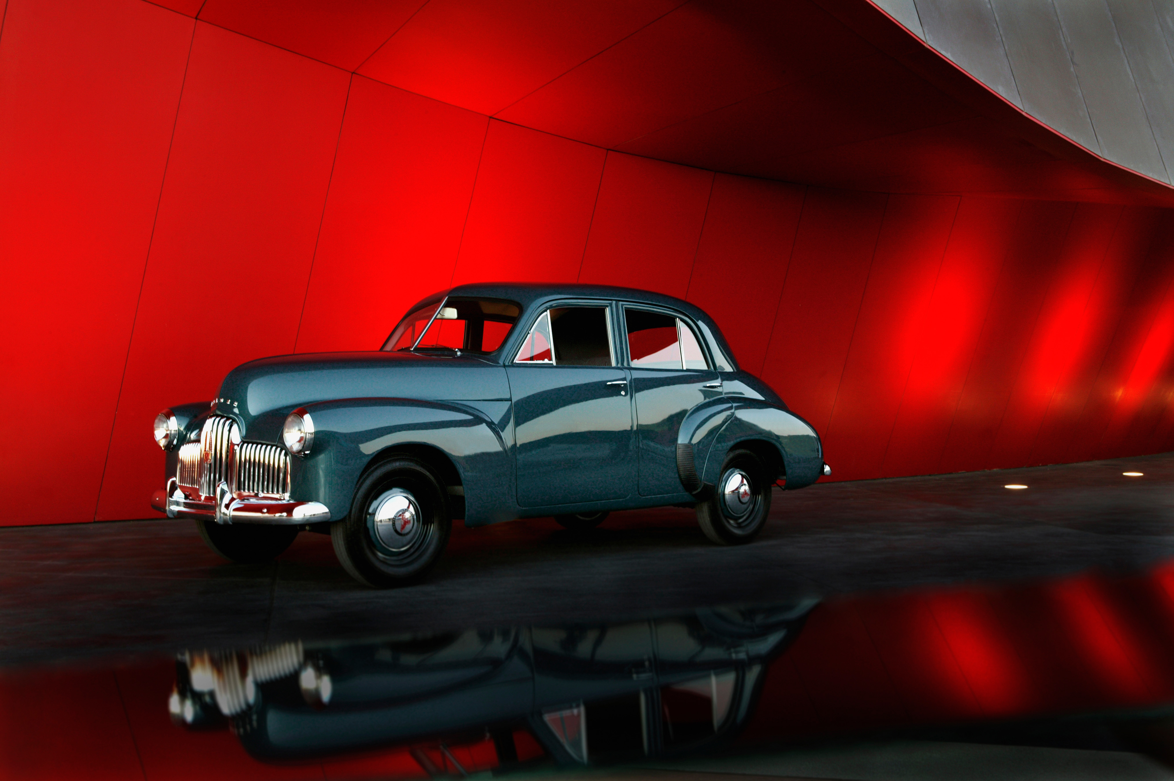 Prototype No.1 Holden sedan (prototype for the 48/215 Model). A 2.15 litre six cylinder, four door, blue-gunmetal grey sedan with chrome-plated radiator grille, bumper bars, and hub caps. The grille and hub caps all bear a single Holden red lion badge, and the lion on the grille badge has a small gold-coloured frame around it. The paint in the centres of the wheels matches the bodywork but the rims are black. There is a vertical division in the middle of the windscreen.