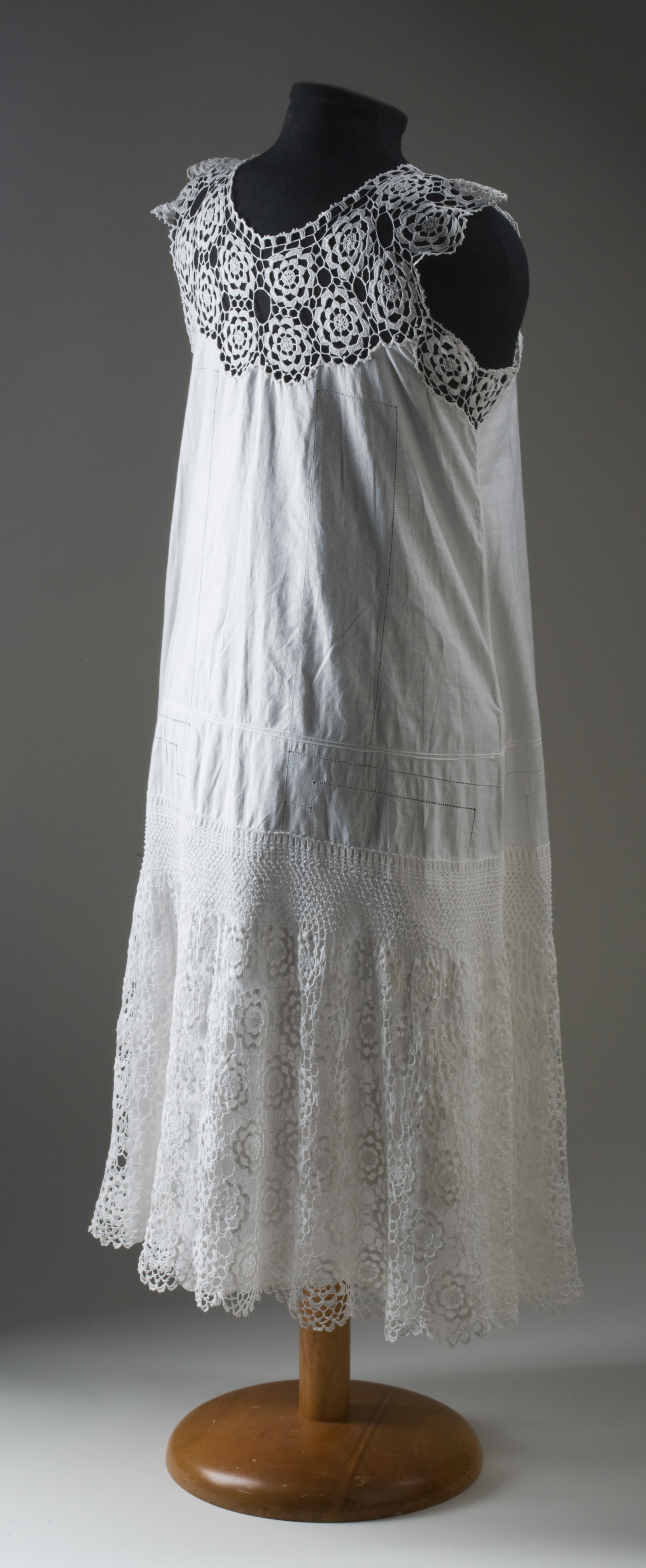  A sleeveless, white, cotton nightdress. The yoke of the nightdress is made from medallion crochet featuring a flower motif. The body of the nightdress is decorated with drawn thread work and the hem features a scalloped edge. A panel of crochet has been placed around the lower part of the nightdress, andf features a straight or plain crochet stitched top below which is medallion crochet in the same pattern as the yoke.
