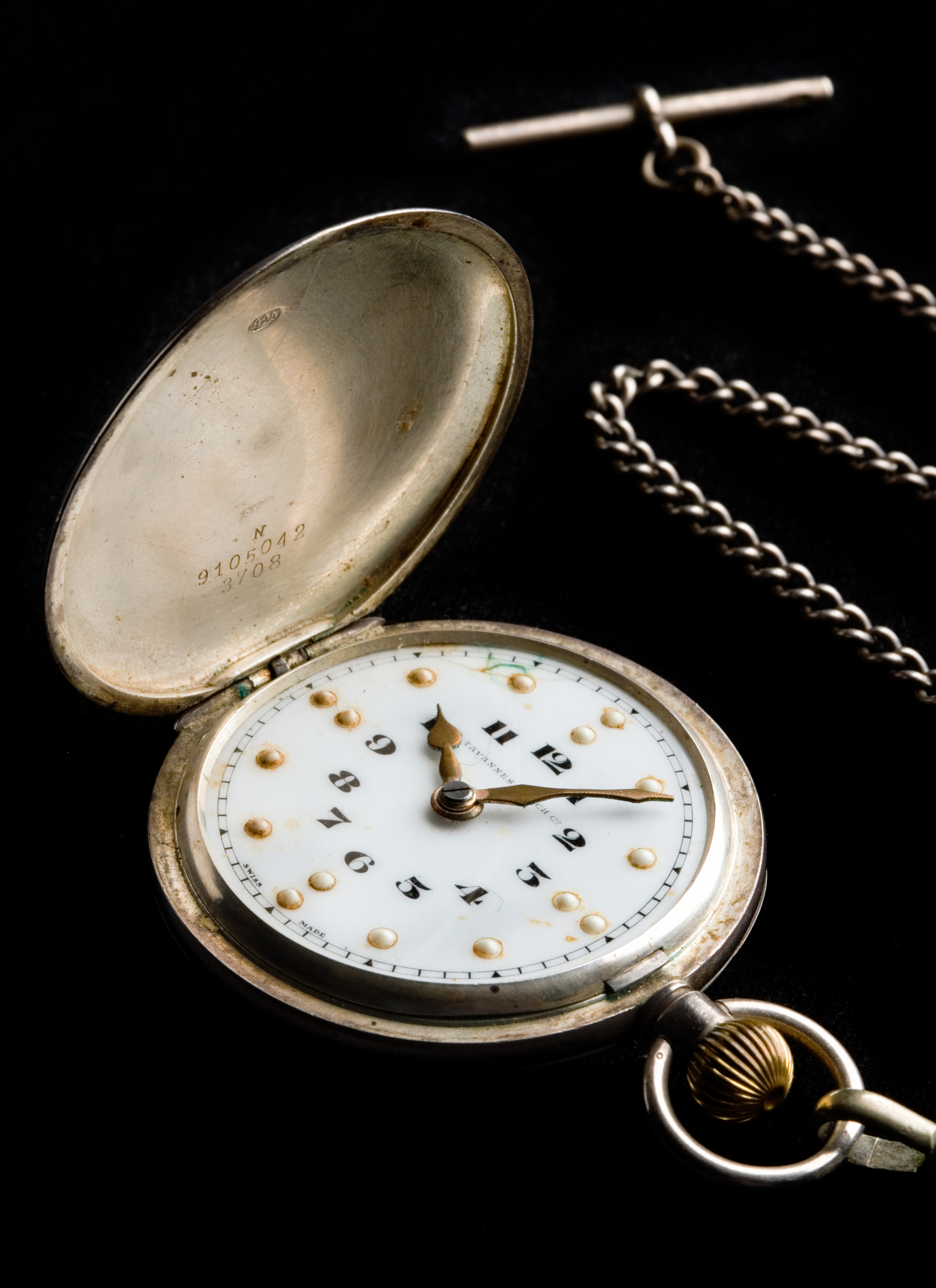 A braille pocket watch which has a black and white enamelled dial with 'TAVANNES WATCH C2' and 'SWISS MADE' lettering, a metal case with a spring-hinged circular lid which is hinged from the left side, and an attached fob chain. The dial has an inner circle of black numbers, a middle circle of raised bumps with two bumps indicating the 12, 3, 6 and 9 positions and single bumps for the remaining. The chain has 'ST S' imprinted on the crossbar, indicating it is made from stainless steel.
