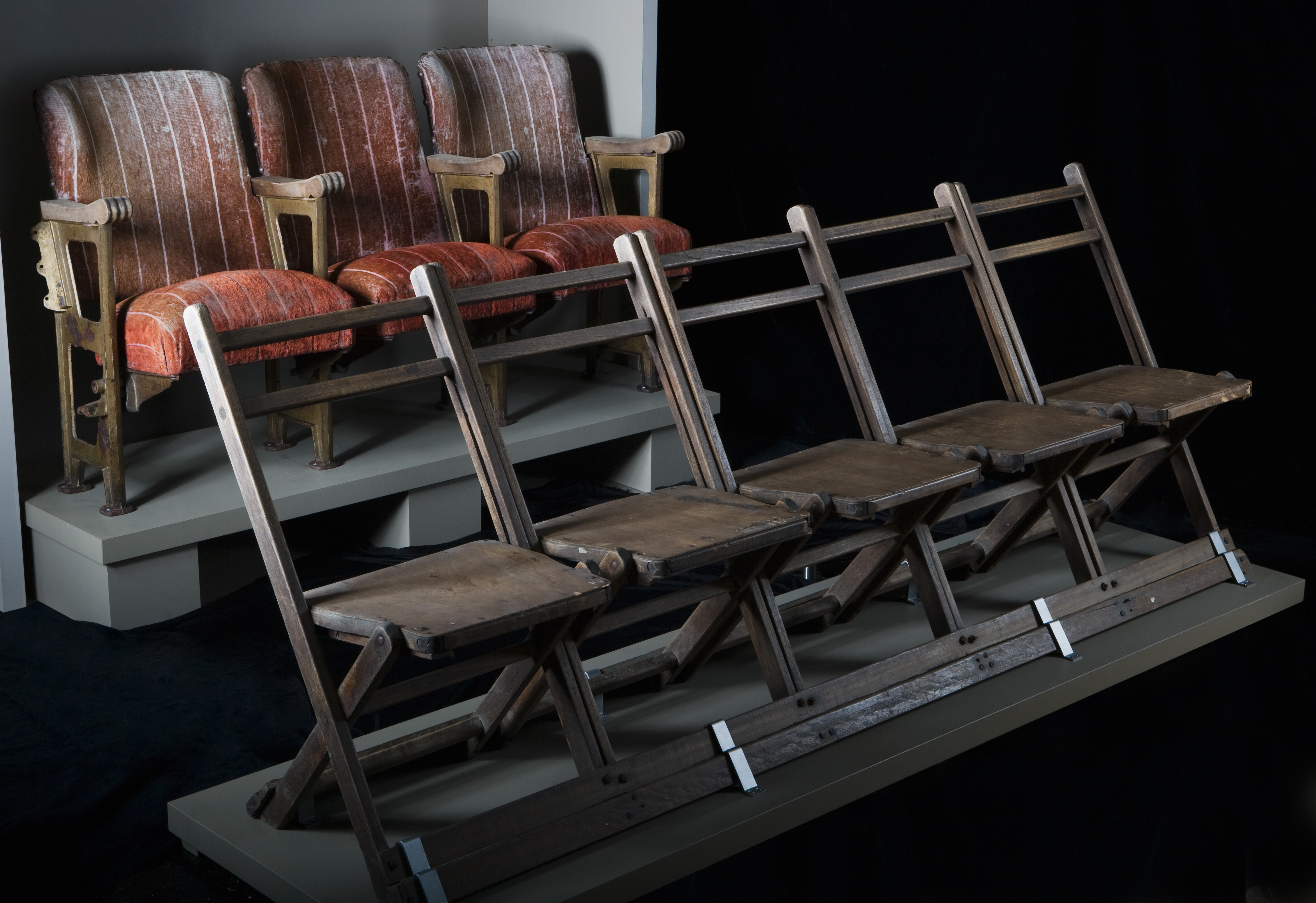  1. A conjoined set of five timber, fold-down theatre seats. The seats feature a flat, solid timber seat and a horizontal timber with two-slat back support. 2. A row of three padded cinema chairs with stripped red plush fabric covering. The backs of the chairs are faded and worn. The chairs are mounted on a solid metal frame which forms the legs of the row. The armrests are made of wood and the base of the seats can be folded up.