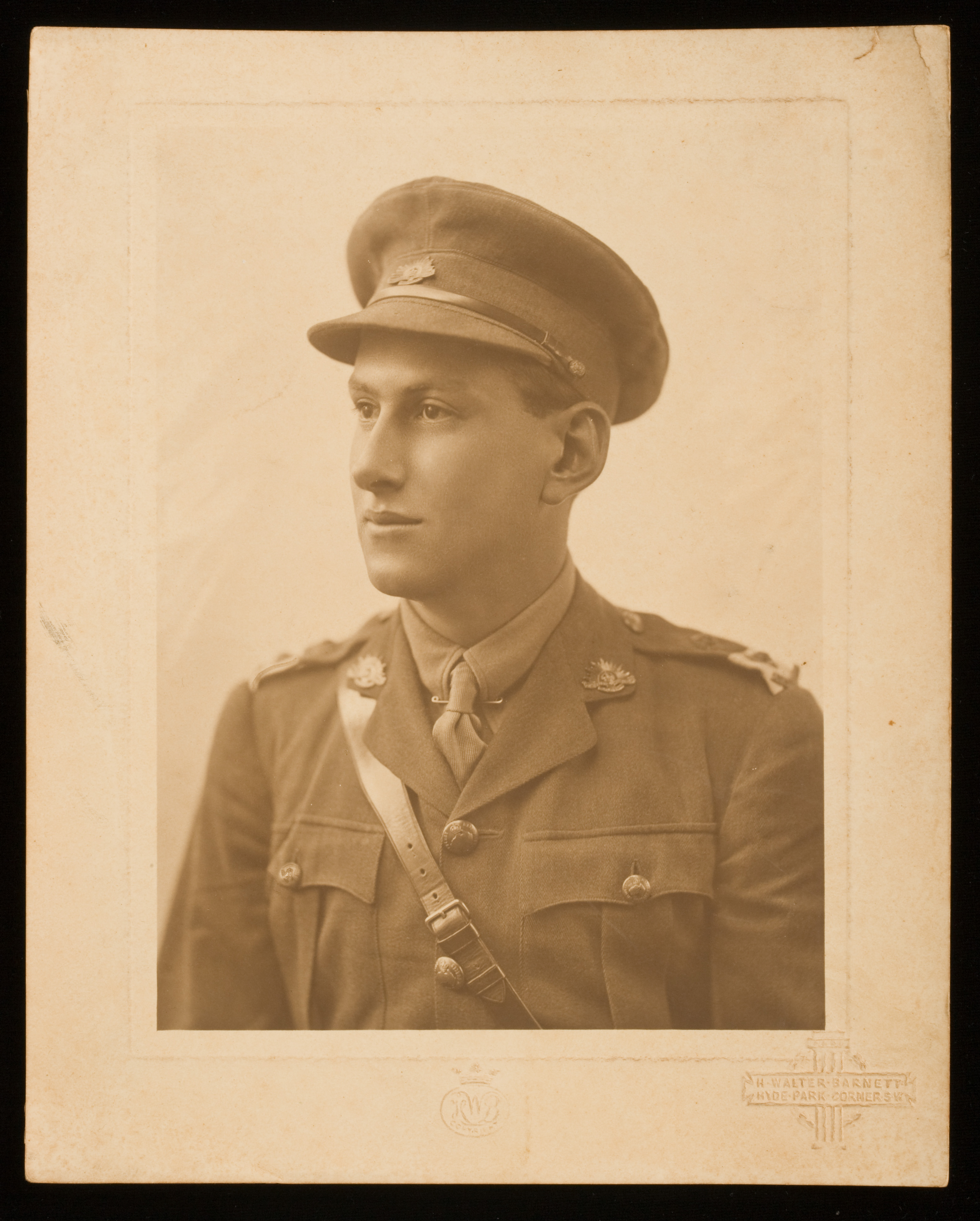  A sepia portrait photograph on light card showing a man, Colyn Cohen, in military uniform. The man wears a peaked cap, a single breasted jacket with pockets, a tie and a leather strap across his torso. His shirt collar is pinned down by one long bar pin, and he wears badges on the jacket collar, epaulettes and hat.