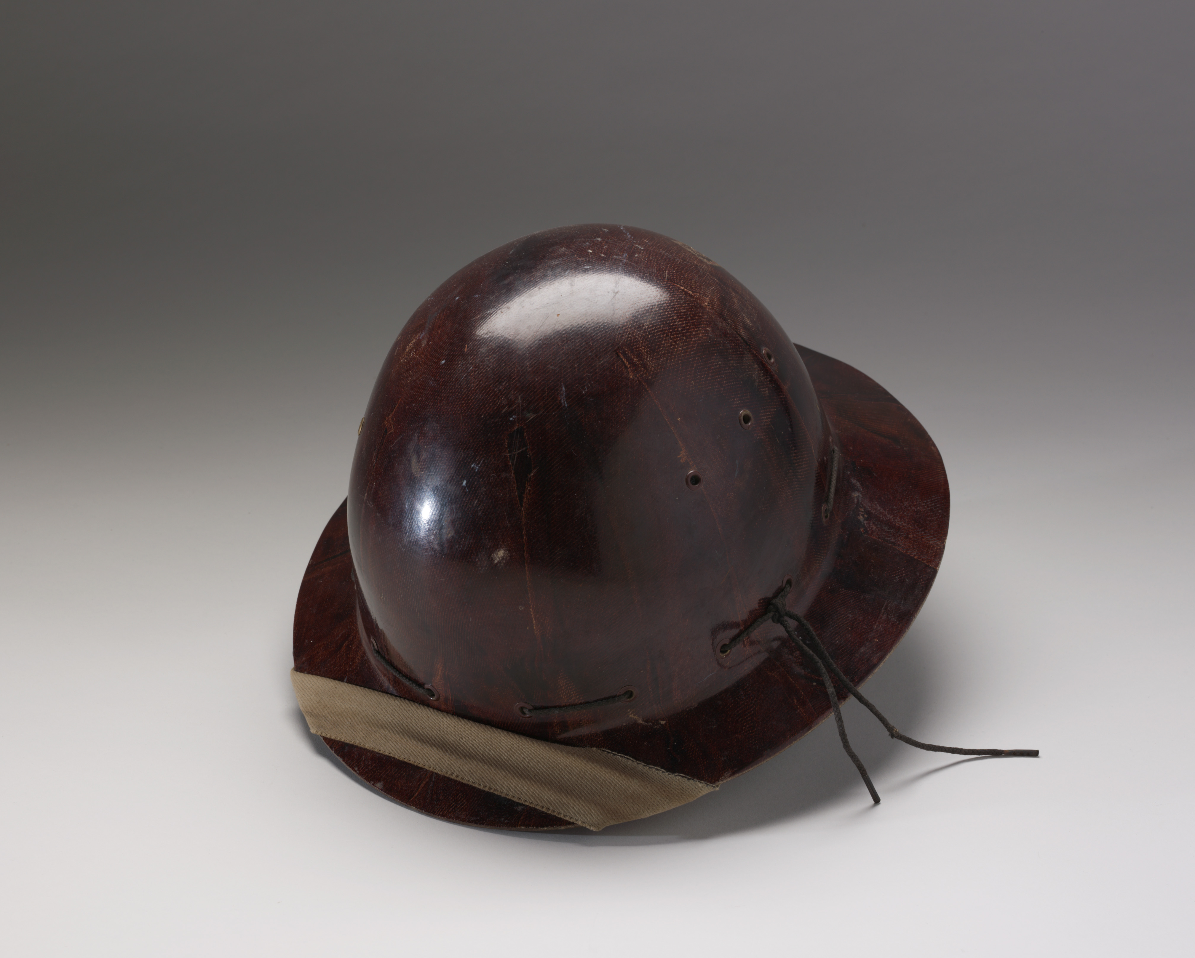  A dark brown hard hat made of Duperite, which is fitted with a khaki webbing chin strap, brown leather headband, and a beige webbing liner. There are scuff/residue marks in the shape of square brackets on the back of the helmet's dome as well as three vent holes on either side.