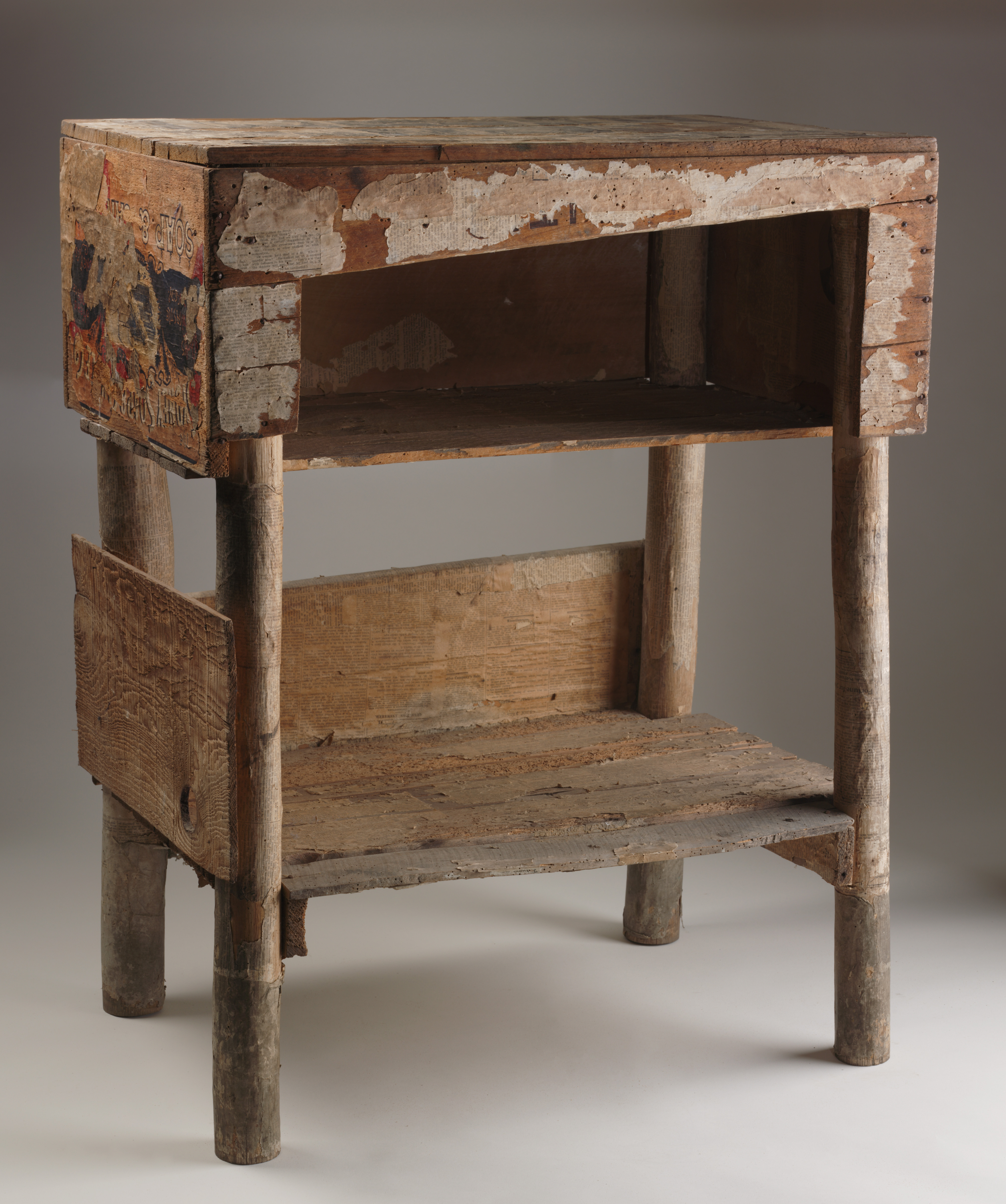  A cupboard made from a pine packing case. The cupboard has two shelves. The top shelf has three walls and a lid, this has been done by removing one side of the pine packing case. The lower shelf has board along two sides. One along the length the other along the width. The cupboard has been covered with newspaper and remnants of this remain. It has four round hardwood legs.