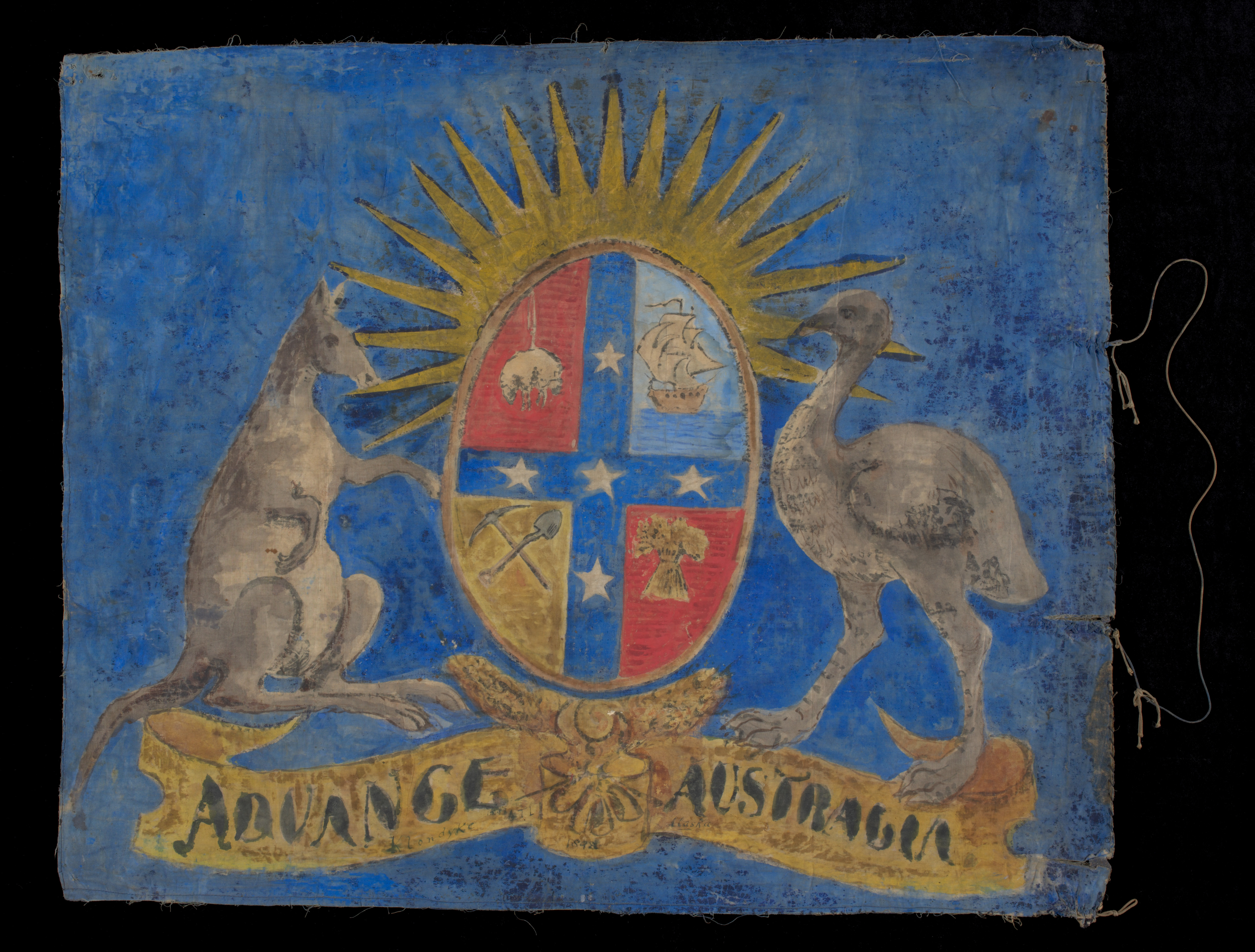  A double sided, hand-painted flag depicting on both sides a kangaroo and an emu either side of a shield, set against a blue background. The shield is broken into four sections showing a sheep, a sailing ship, a sheaf of wheat and a pick and shovel. The stars of the Southern Cross separate the quadrants and a rising sun appears above the shield. A banner below the coat of arms reads 'ADVANCE AUSTRALIA'. There are distinct differences on either side of the flag, particularly in the representations of the kan